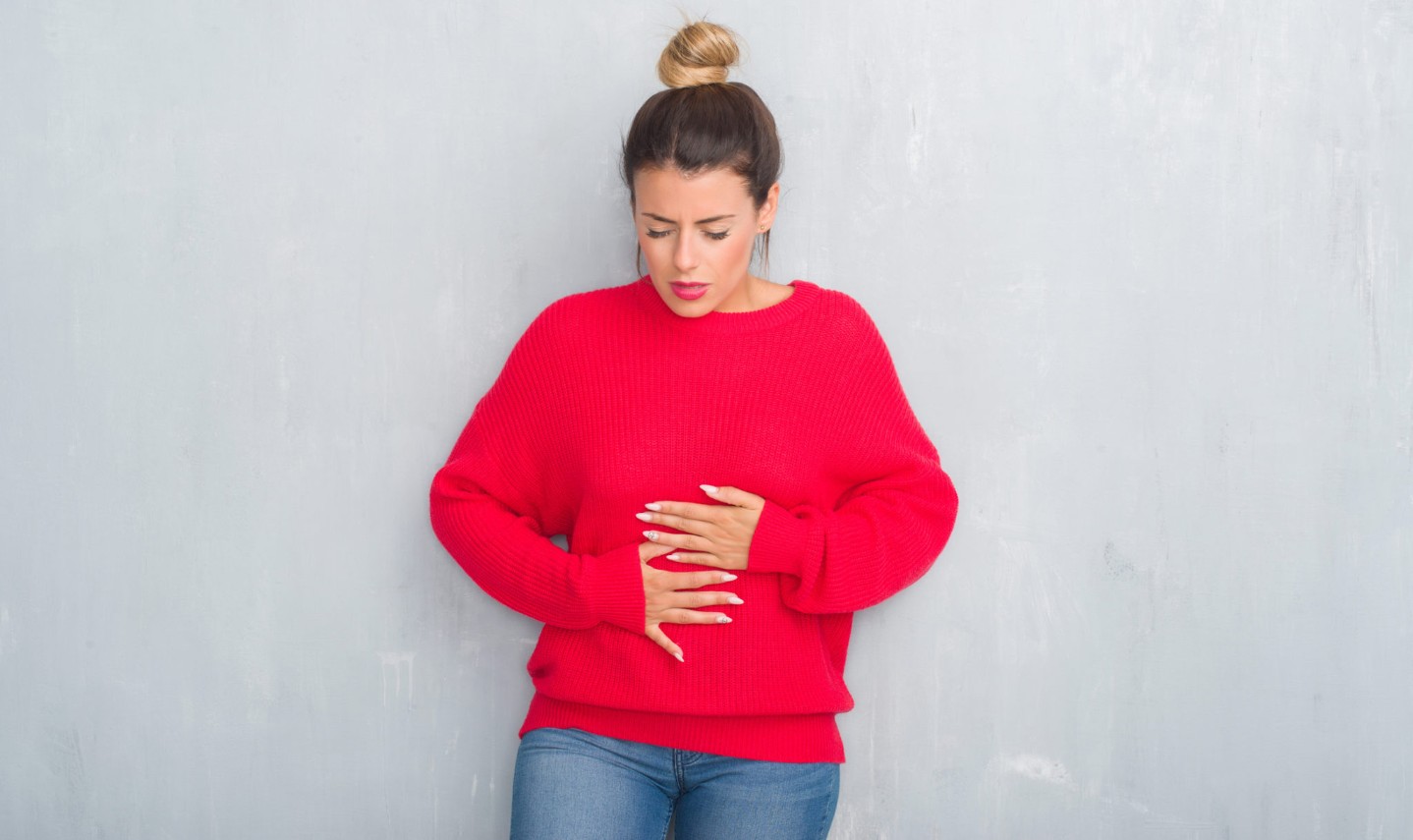 Young adult woman over grey grunge wall wearing winter outfit with hand on stomach because indigestion, painful illness feeling unwell.||stomach bloating