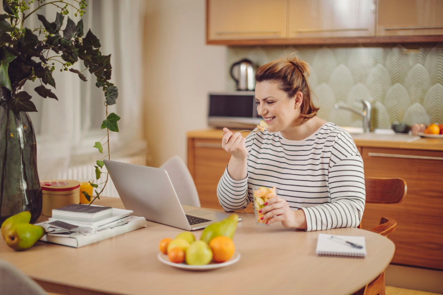 Young woman at home eating healthy fruit and working on her laptop
