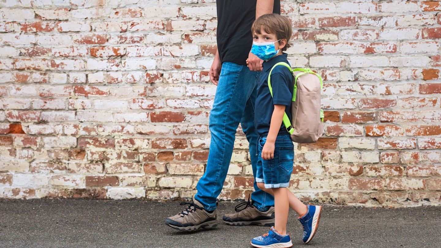 child in a mask with a backpack holding a parent's hand